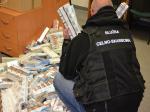 Na zdjęciu umundurowany funkcjonariusz Służby Celno-Skarbowej przed połozonymi na popdłodze paczkami papierosów. W ręku trzyma dwa kartony z papierosami.