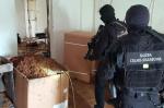 Umundurowani funkcjonariusze Służby Celno-Skarbowej w pomieszczeniu nielegalnej krajalni tytoniu oglądają pudła z nielegalnym tytoniem.