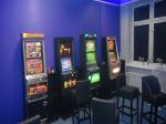 Cztery automaty do gier hazardowych stoją pod ścianą, przed nimi stoją cztery wysokie krzesła barowe.  