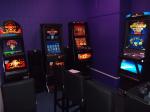 Trzy automaty do gier hazardowych stoją pod jedną ścianą, pod ścianą obok stoi kolejny automat. Przed automatami stoją wysokie krzesła barowe.  
