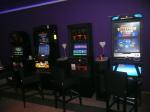 Cztery automaty hazardowe stojące pod ścianą. Przed nimi stoją wysokie stołki barowe.